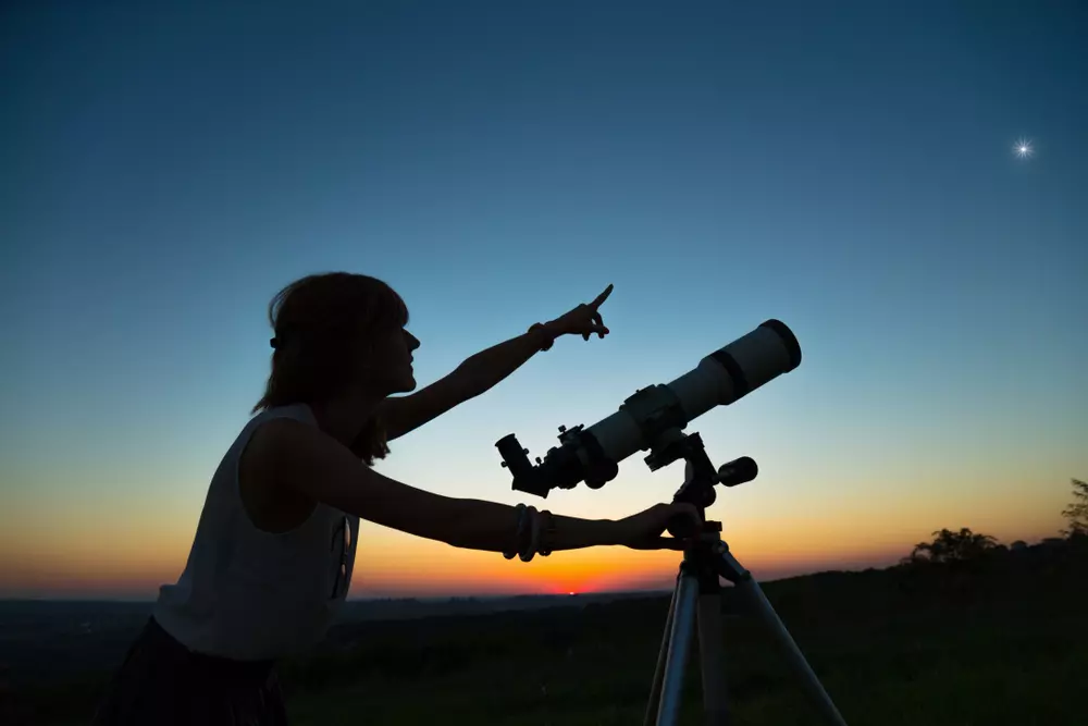 Mulher utilizando um telescópio na natureza ao entardecer, simbolizando a conexão entre ciência e conhecimento indígena para compreender quem não 'fala' como nós.