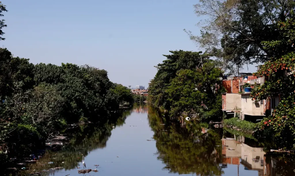 Um rio urbano passando por uma comunidade vulnerável, com moradias precárias ao redor e vegetação densa, demonstrando desigualdade e vulnerabilidade urbanas.