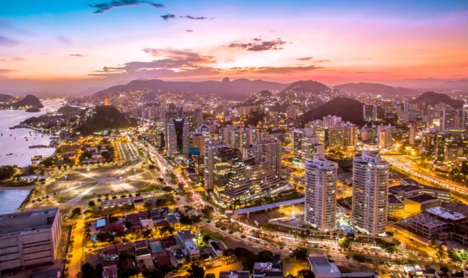 Vista panorâmica da cidade do Rio de Janeiro ao entardecer, com luzes iluminando os prédios, montanhas ao fundo e o céu colorido ao pôr do sol.