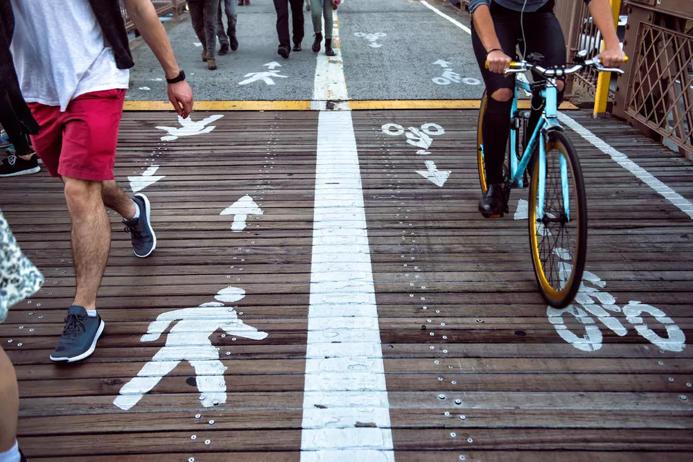 Benefícios do The Big U para a cidade, com ciclovia e espaço para pedestres em ambiente urbano. Pessoas usando bicicletas e caminhando em uma rua com infraestrutura sustentável.