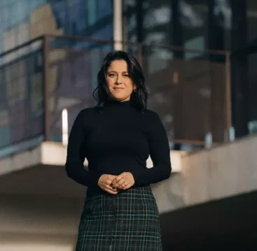 Fotografia de Ana Carolina Vaz em frente a uma construção moderna, vestindo blusa preta e saia de xadrez, com expressão confiante e cenário urbano ao fundo.