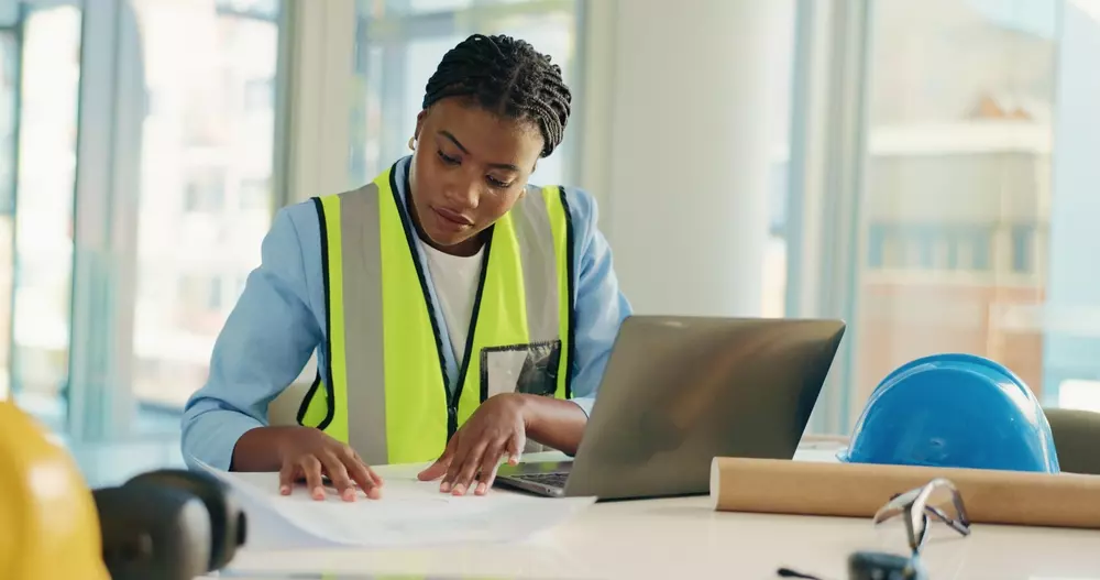 Engenheira de construção avalia projetos de trabalho em escritório com laptop e capacete de segurança na mesa