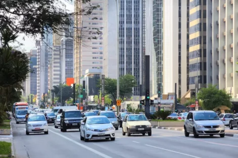 uma rua movimentada em uma cidade moderna, com veículos diversos, árvores e prédios altos ao fundo, evidenciando o streetscape