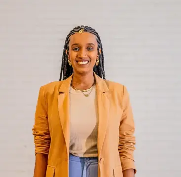 Audrey Carolini, arquiteta, urbanista e designer, posando com sorriso e vestindo blazer bege ao lado de parede neutra