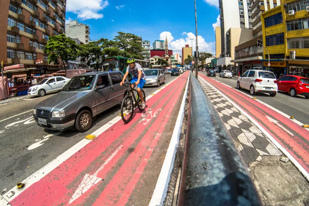 Ciclovia urbana em cidade movimentada com ciclista pedalando ao lado de carros, edifícios altos e céu azul, promovendo mobilidade sustentável e segurança para ciclistas.