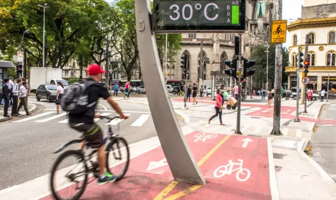 Ciclovia ativa em uma rua movimentada de uma cidade, com ciclistas, pedestres, sinalizações e clima de calor de 30°C, destacando a pegada ambiental da IA na mobilidade sustentável.