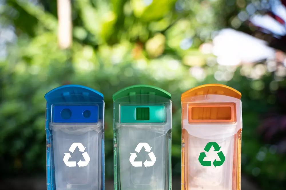 Três lixeiras de coleta seletiva para reciclagem de resíduos recicláveis, coloridas em azul, verde e laranja, com fundo natural e árvore ao fundo, promovendo a sustentabilidade e a separação adequada do lixo.