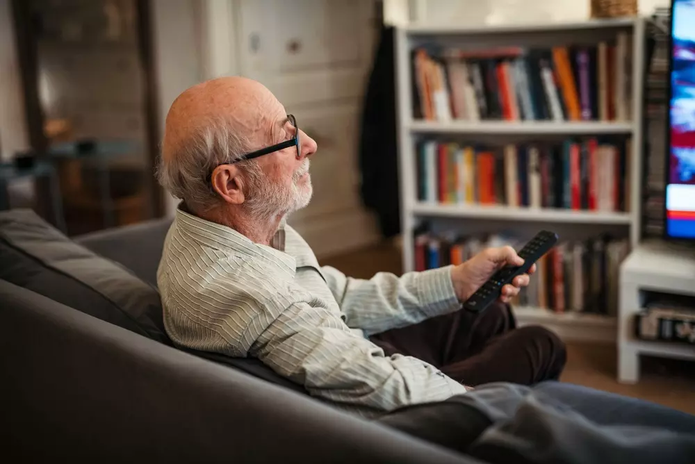 	
Homem idoso sentado no sofá assistindo TV com controle remoto na mão, ao fundo uma estante de livros em um ambiente de sala de estar.